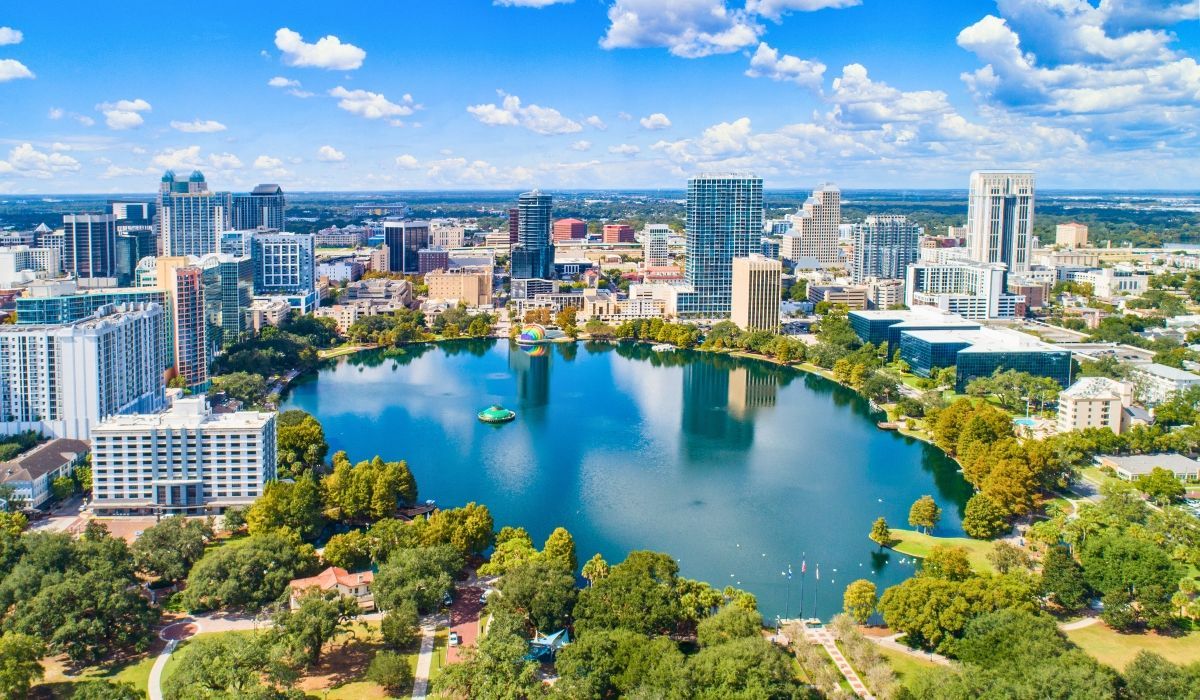 A panoramic view of downtown Orlando, Florida, featuring modern high-rise buildings, Lake Eola Park, and surrounding greenery under a clear blue sky.