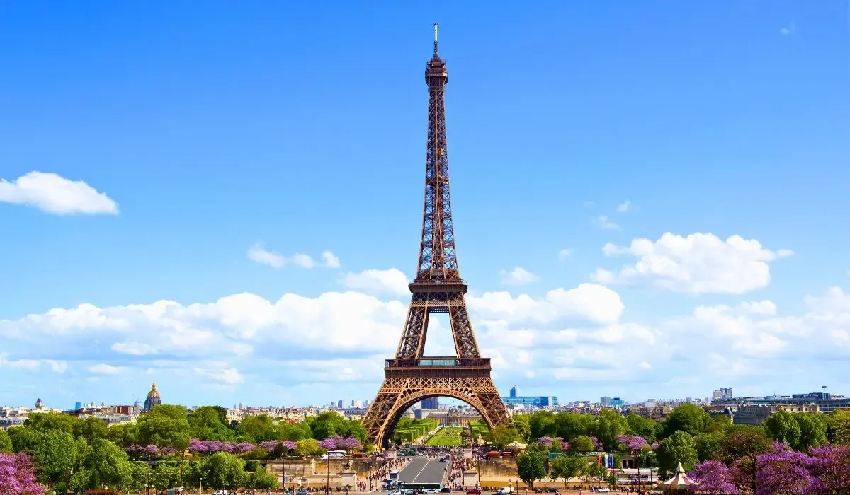 Iconic Eiffel Tower rising over Paris, France, under a clear blue sky surrounded by green trees and blooming flowers. Canadians considering property purchases in France can access competitive CAD to EUR rates and cost-saving transfers with MTFX.