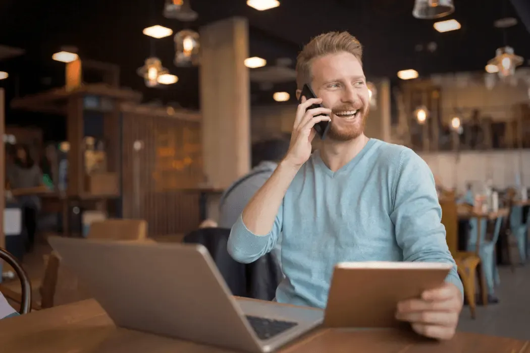 Smiling man working remotely at a café, talking on the phone while using a tablet and laptop.