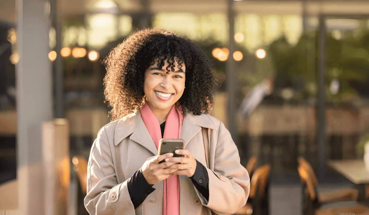 Smiling woman using her smartphone to send money internationally, standing outside a modern café with warm lighting in the background.