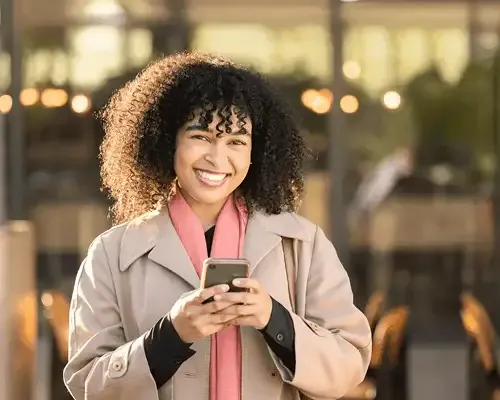 Smiling woman using her smartphone to send money internationally, standing outside a modern café with warm lighting in the background.