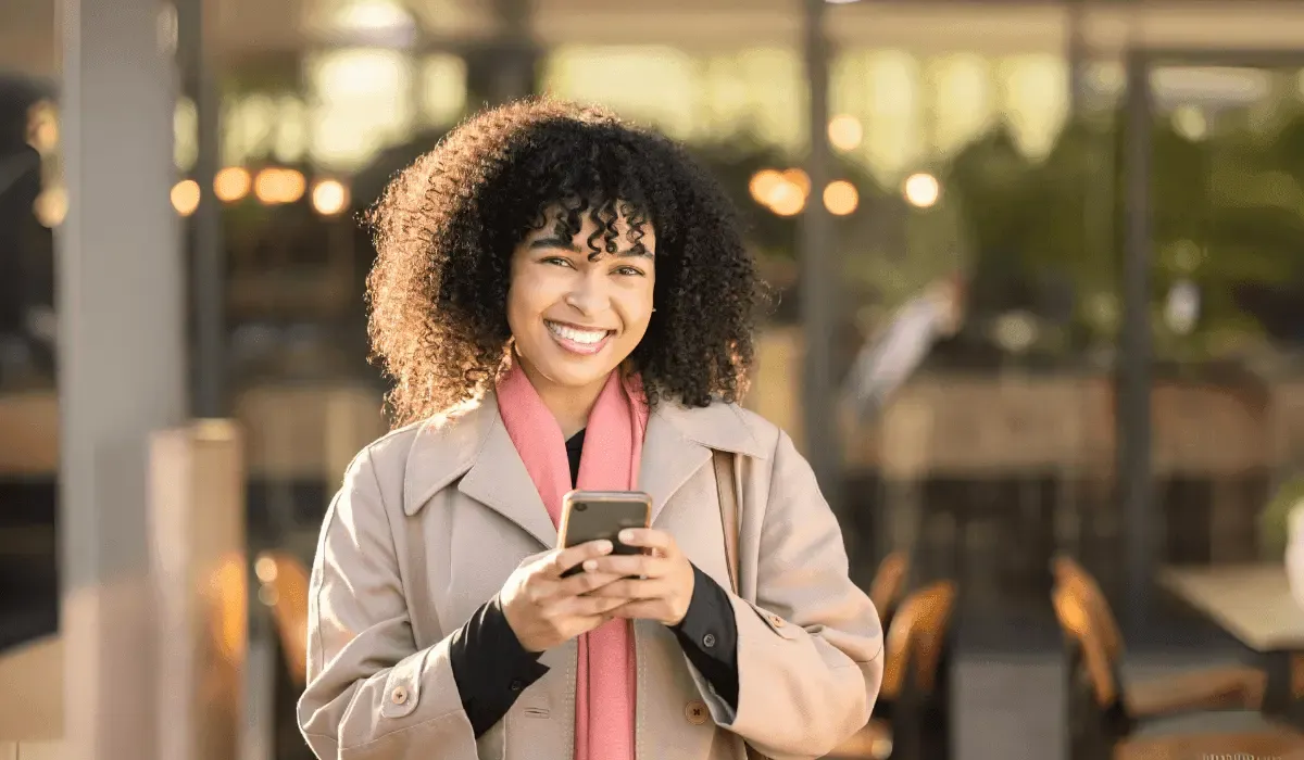 Smiling woman using her smartphone to send money internationally, standing outside a modern café with warm lighting in the background.