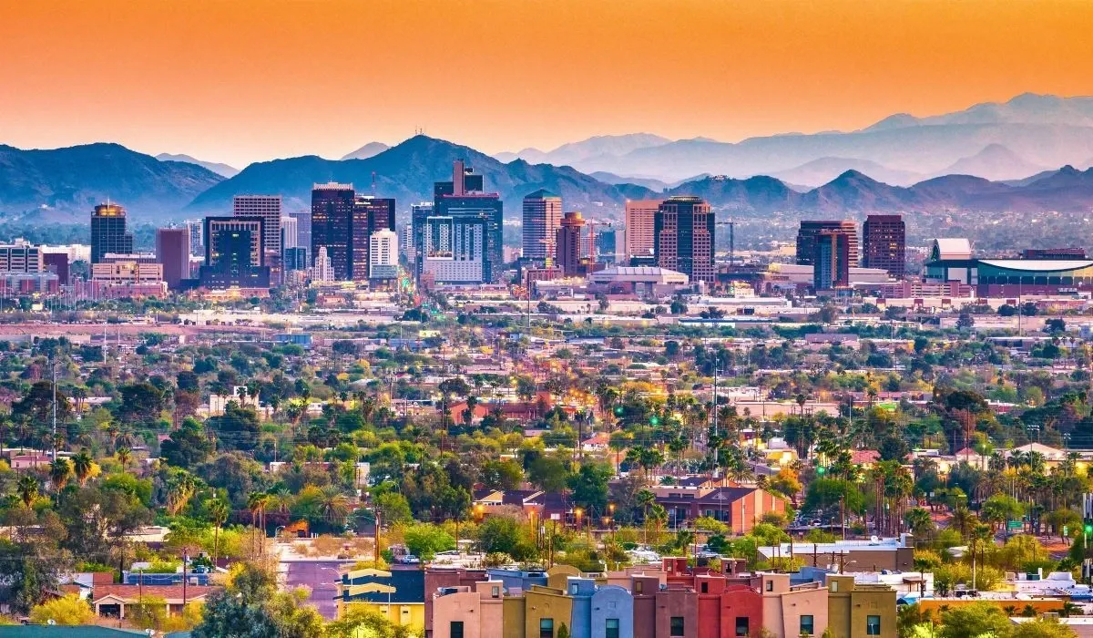 Panoramic cityscape of Phoenix, Arizona at sunset with downtown buildings, desert terrain, and distant mountain ranges.