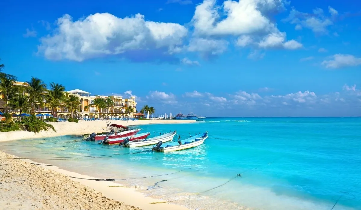 A tranquil Caribbean beach in Playa del Carmen, Mexico, featuring anchored boats, palm trees, and a luxury beachfront resort.