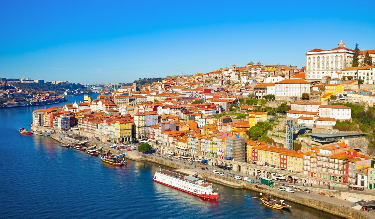 A lively riverside scene in Porto with traditional boats, colorful architecture, and red-tiled rooftops.