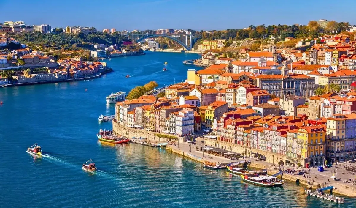 Colourful waterfront buildings along the Douro River in Porto, Portugal, with traditional boats, blue water and a scenic bridge in the background on a sunny day.