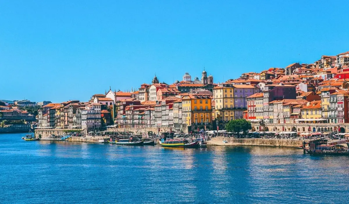 A panoramic view of Lisbon, Portugal, featuring colorful buildings, the Tagus River, and the iconic 25 de Abril Bridge under a clear sky.
