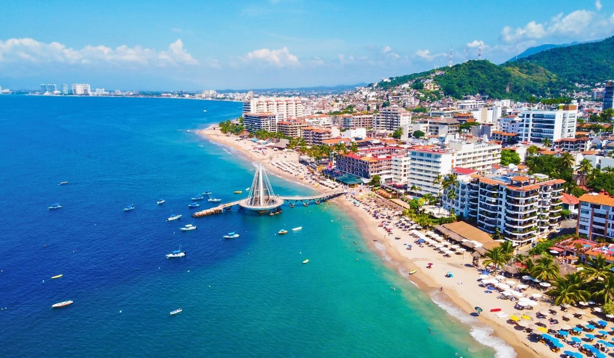 An aerial shot of Puerto Vallarta's coastal cityscape, featuring the Malecón boardwalk, historic architecture, and surrounding mountains.