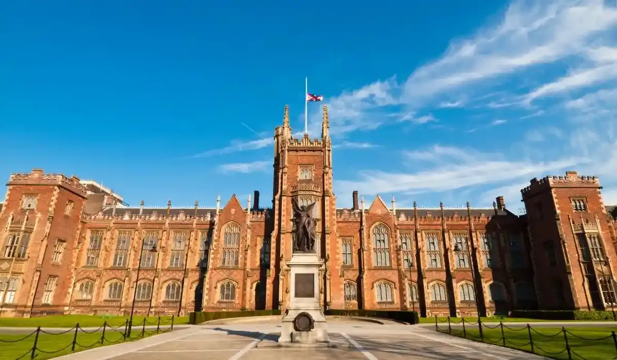 Front view of Queen’s University Belfast with red-brick Gothic architecture, a central tower, and a statue in the foreground under a blue sky.