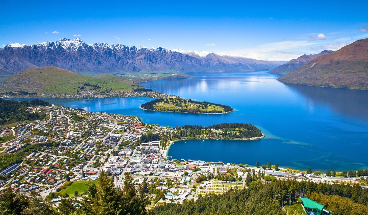 A picturesque aerial view of Queenstown in New Zealand showing urban residential area with lots of lush greenery and trees and mountains and ocean in the backdrop.
