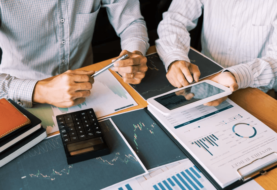 Two business professionals reviewing financial charts and market data on a tablet and printed reports, with a calculator and graphs on the desk during a financial analysis meeting.