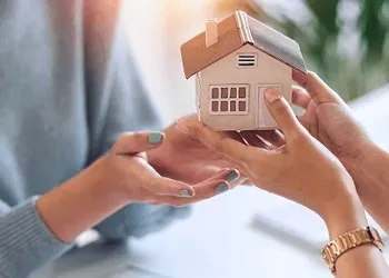 Hands exchanging a small house model, representing receiving rental income or managing property earnings.