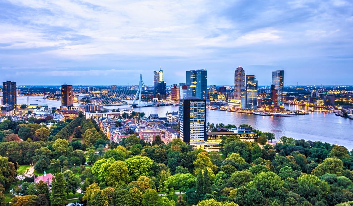 Rotterdam skyline at sunset with modern skyscrapers, the iconic Erasmus Bridge, and lush greenery in the foreground.