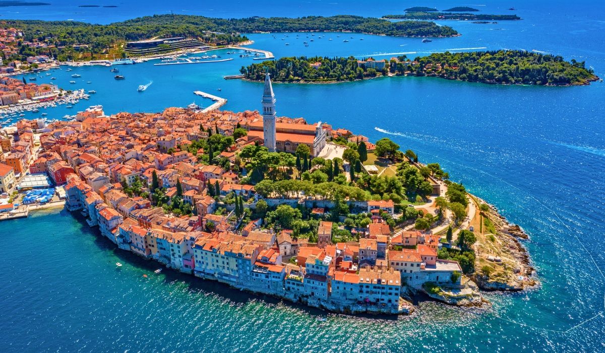 Aerial shot of Rovinj's peninsula with tightly packed red-roofed buildings and a prominent church tower surrounded by turquoise waters.