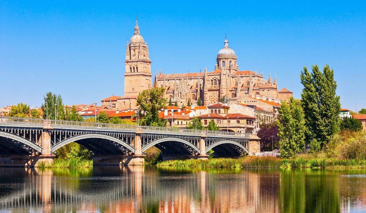 A view of the New Cathedral of Salamanca with its detailed Gothic and Baroque architecture, rising above the city’s rooftops, with a stone bridge spanning the Tormes River in the foreground.