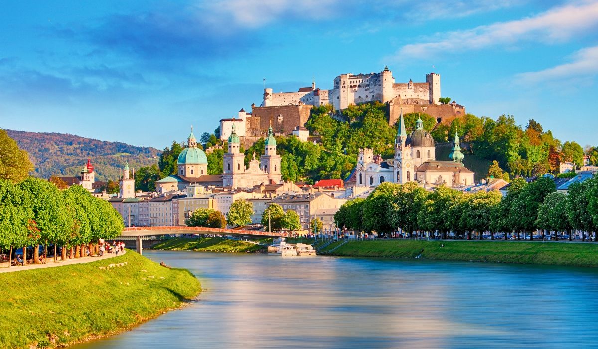 A panoramic view of Salzburg, Austria, with the Hohensalzburg Fortress overlooking baroque architecture and the Salzach River under a clear sky.
