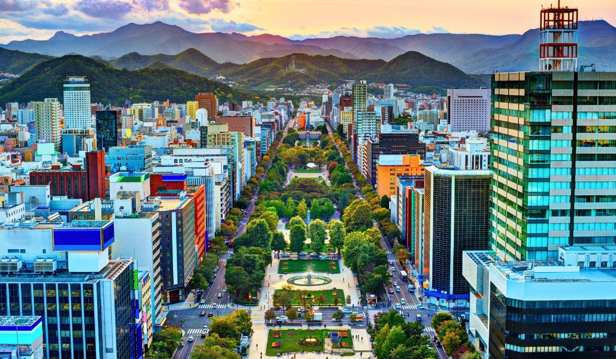 Aerial view of Sapporo’s Odori Park lined with trees and surrounded by tall buildings, backed by mountain ranges at sunset.