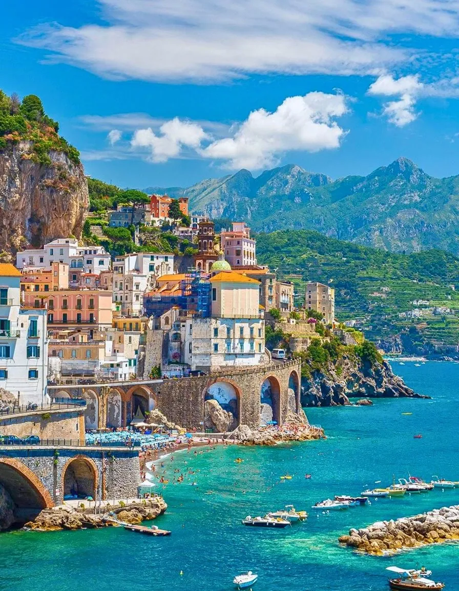 Scenic coastal town in Italy with colorful cliffside buildings, turquoise waters, boats, and mountains in the background