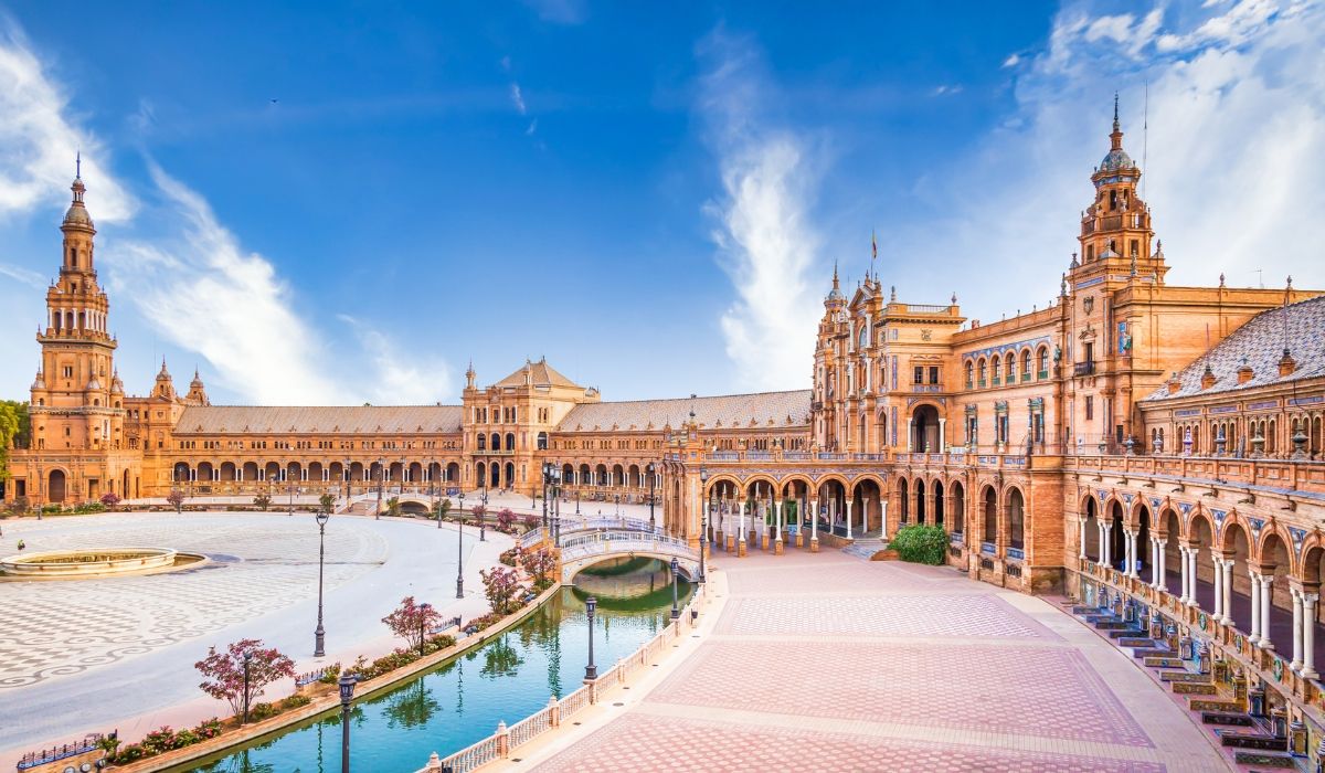 The grand Plaza de España in Seville, featuring its curved Renaissance Revival-style building, arched bridges over a canal, and a large tiled central plaza under a vivid blue sky.