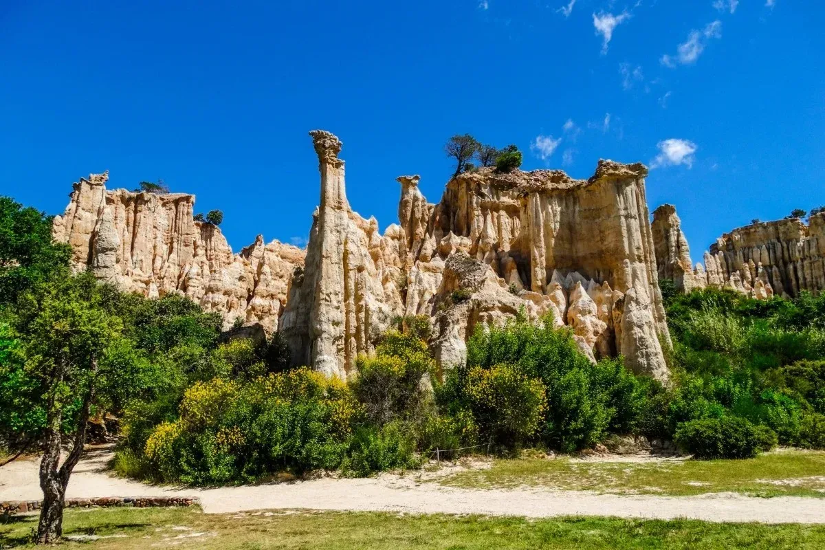 Dramatic pale rock cliffs and tall eroded stone formations rise above green shrubs and trees beneath a clear blue sky.