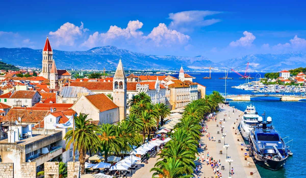 A vibrant view of Split's historic Riva waterfront, featuring palm trees, yachts, and the bell tower of the Cathedral of Saint Domnius set against a mountainous backdrop.