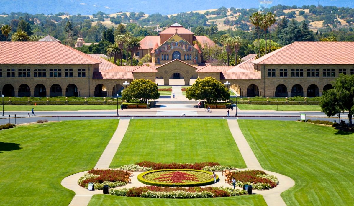 Stanford University's Main Quad with red-tiled Spanish Revival buildings, symmetrical gardens, and rolling California hills in the background.