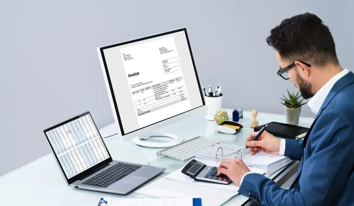 A picture of a man in a professional setting reviewing financial documents, with a computer displaying an invoice, a laptop showing financial data, and a calculator on the desk.