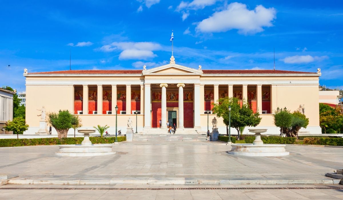 A grand neoclassical building with columns, statues, and a Greek flag, representing the National and Kapodistrian University of Athens on a sunny day.