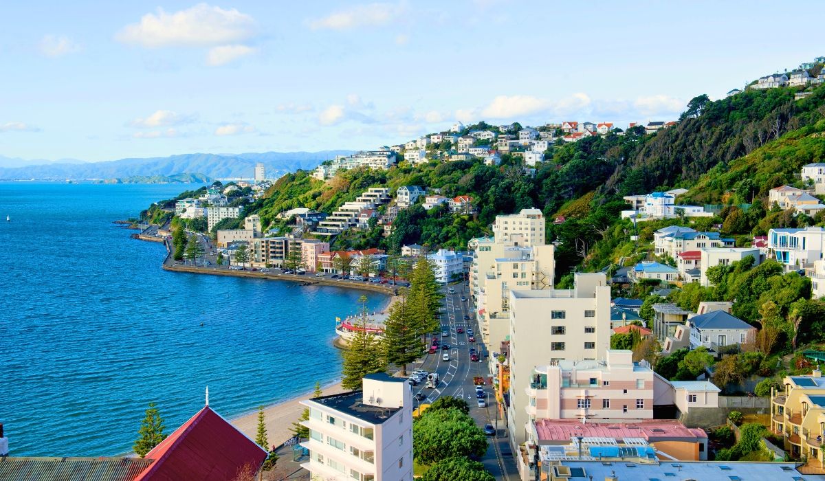 A picturesque coastal city with hillside homes, a winding road, and sparkling blue waters on a sunny day.