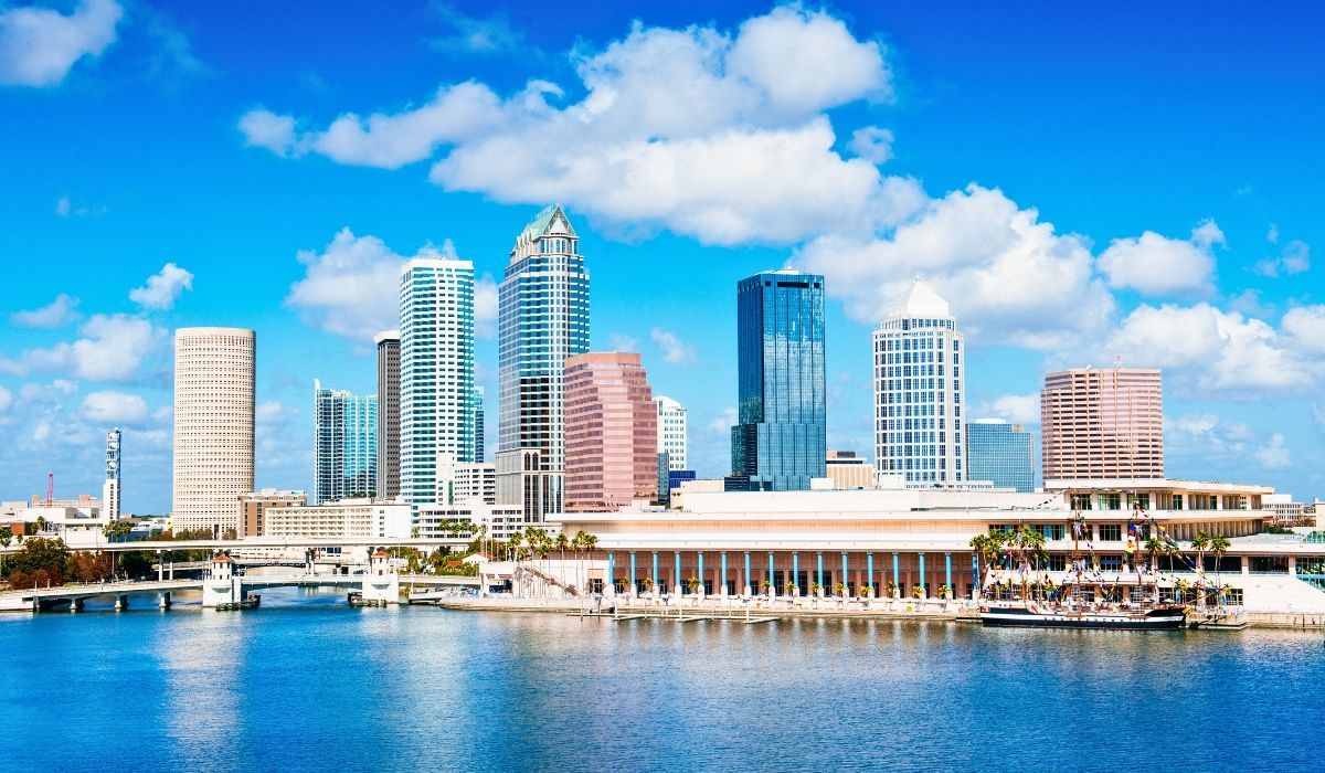 An aerial view of downtown Tampa, Florida, displaying a mix of modern skyscrapers, the Hillsborough River, and the city's waterfront areas under a partly cloudy sky.