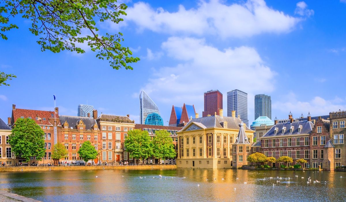 Historic buildings of The Hague reflected in a pond, with modern skyscrapers rising in the background under a bright blue sky.
