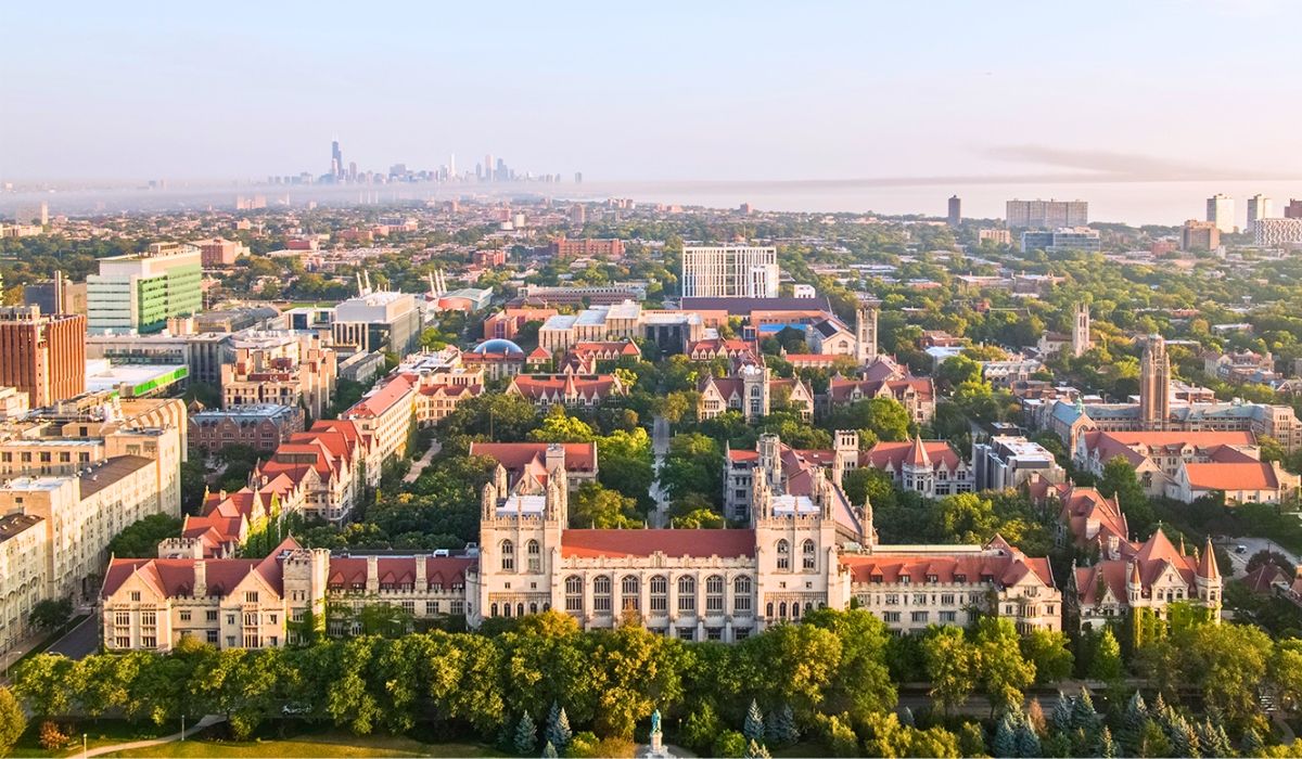 Expansive aerial view of the University of Chicago campus featuring Gothic-style buildings, tree-lined streets, and the distant Chicago skyline under hazy skies.