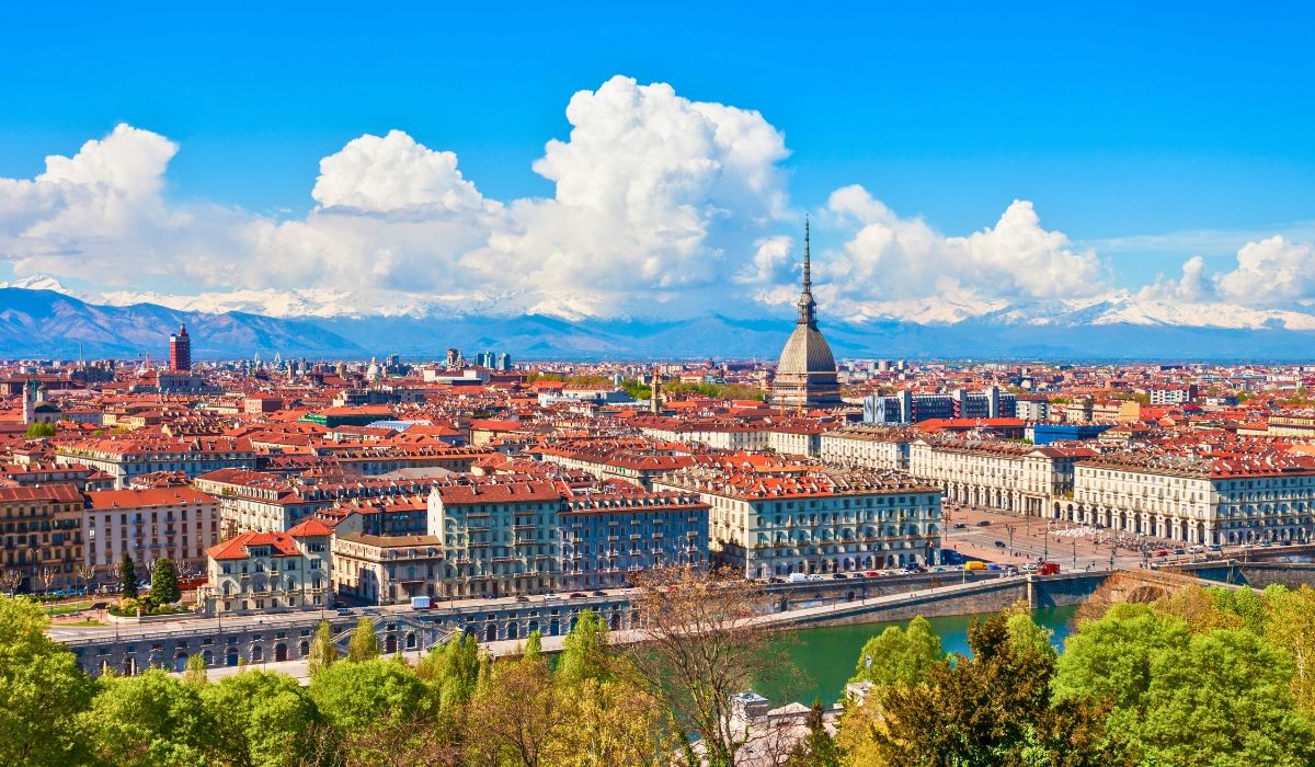 An aerial shot of Turin, highlighting the Mole Antonelliana towering above the city's rooftops, with the Alps in the background.