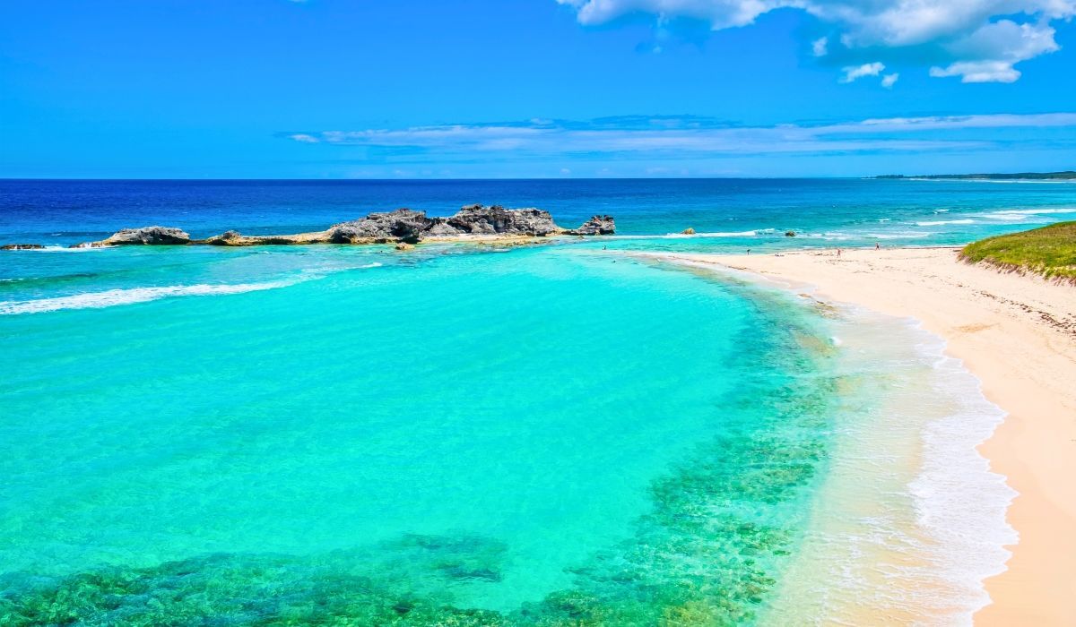 Pristine beach in Turks and Caicos with crystal-clear turquoise waters, coral reef patches, and soft white sand under a bright blue sky.