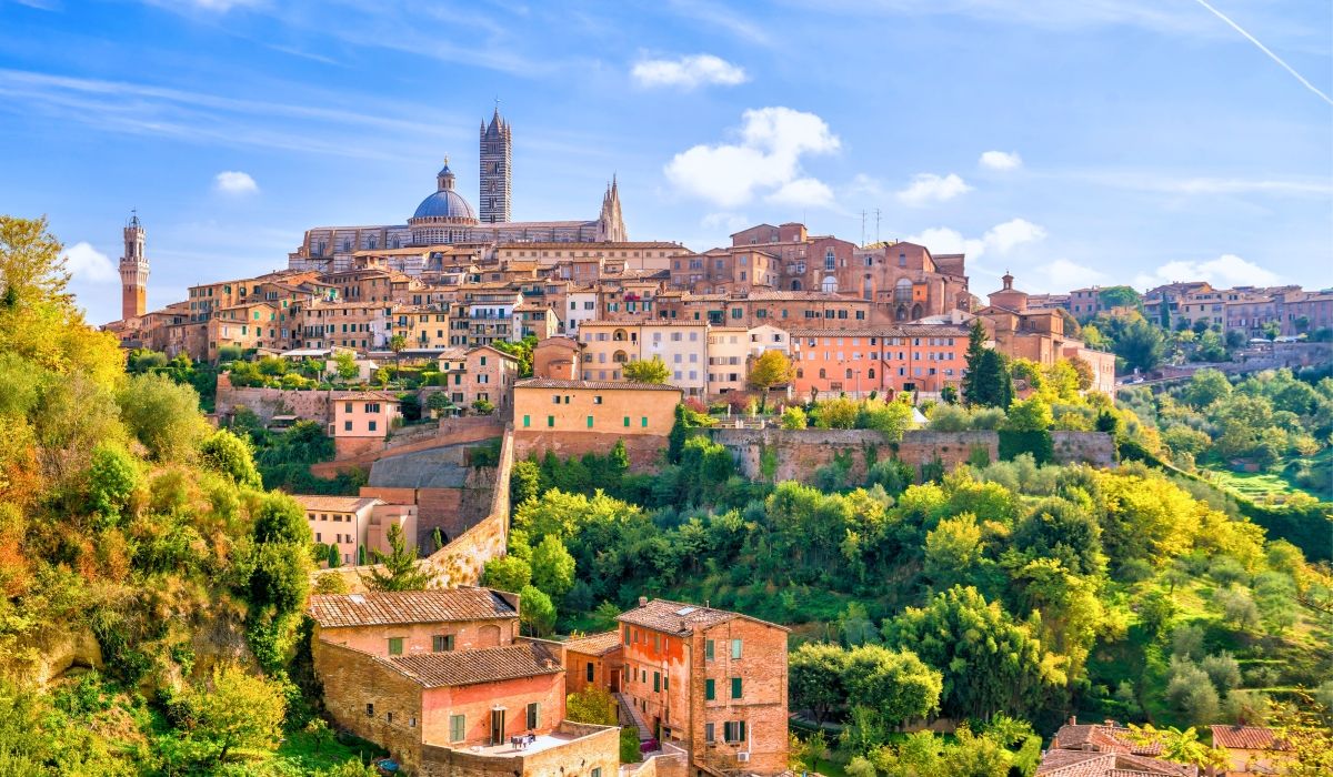 The medieval skyline of Siena, Italy, with terracotta buildings and historic towers atop lush green hills.