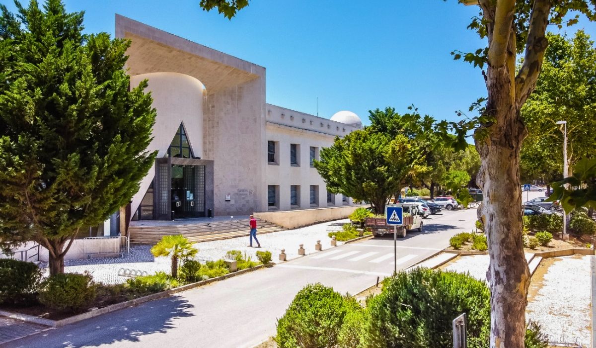 A coastal view of Universidade do Algarve, showing campus buildings near the shoreline with the ocean visible in the distance.