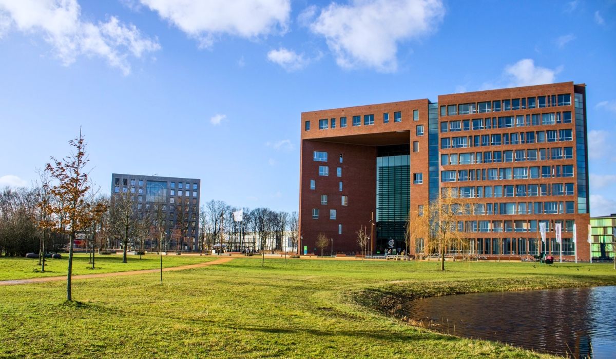 A modern academic building on the Wageningen University & Research campus in the Netherlands, featuring a red-brick structure with large windows and open green space in the foreground under a blue sky.