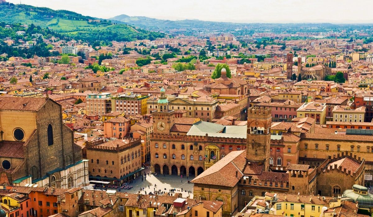 Aerial view of Bologna, Italy, showcasing terracotta rooftops, historic towers, and the main square of Piazza Maggiore.