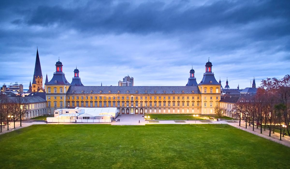 Illuminated evening view of the University of Bonn's main yellow baroque-style building with a wide lawn in the foreground and church towers in the background.