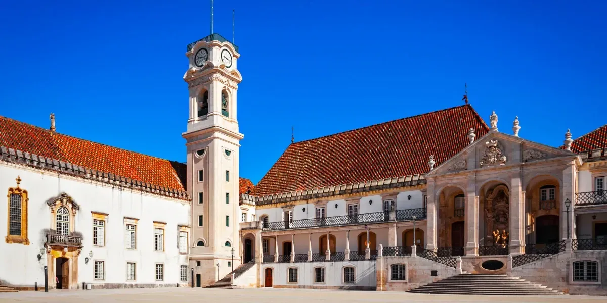 Historic University of Coimbra building in Portugal with a clock tower, red-tiled roofs, and a clear blue sky.