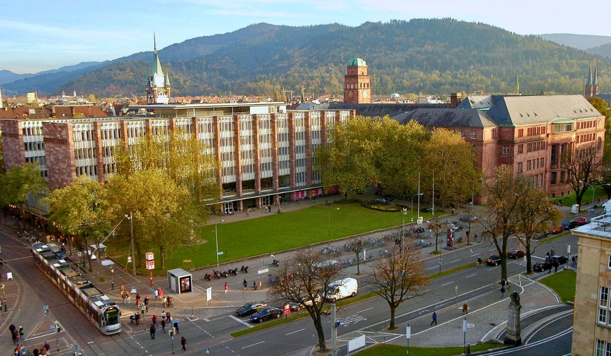 Aerial view of the University of Freiburg's campus with modern and historic buildings, green lawns, and the Black Forest mountains in the background.