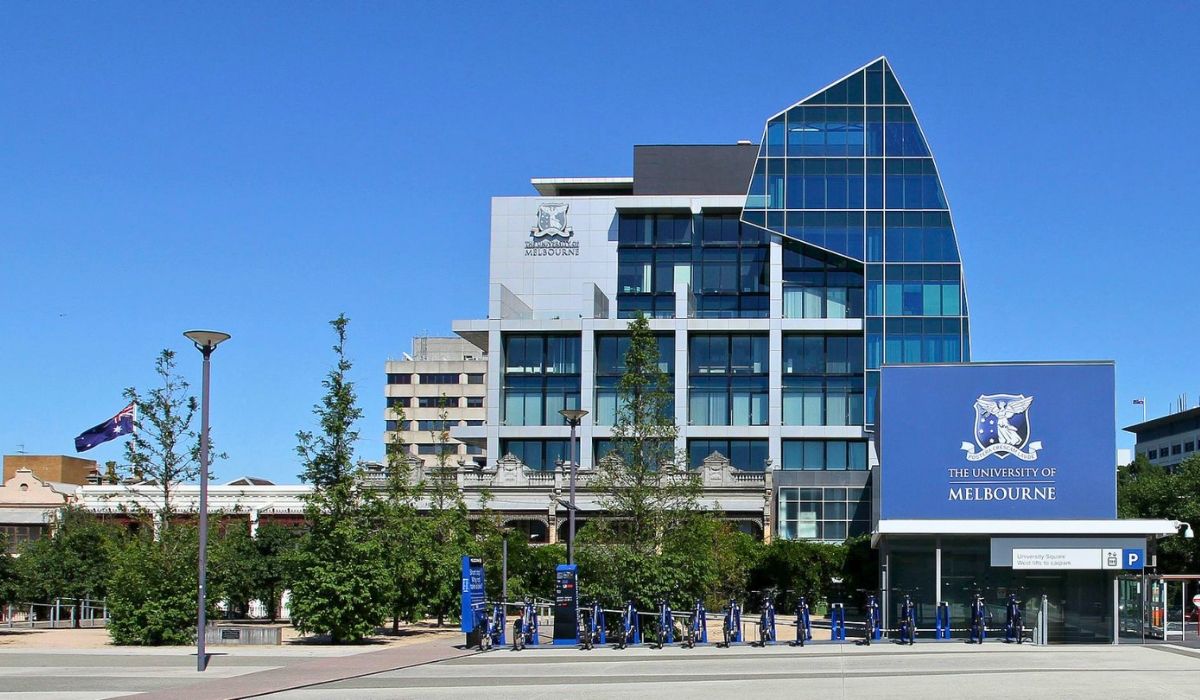 University of Melbourne’s main campus featuring a modern glass building, large signage, bike racks, and Australian flags under a clear blue sky.