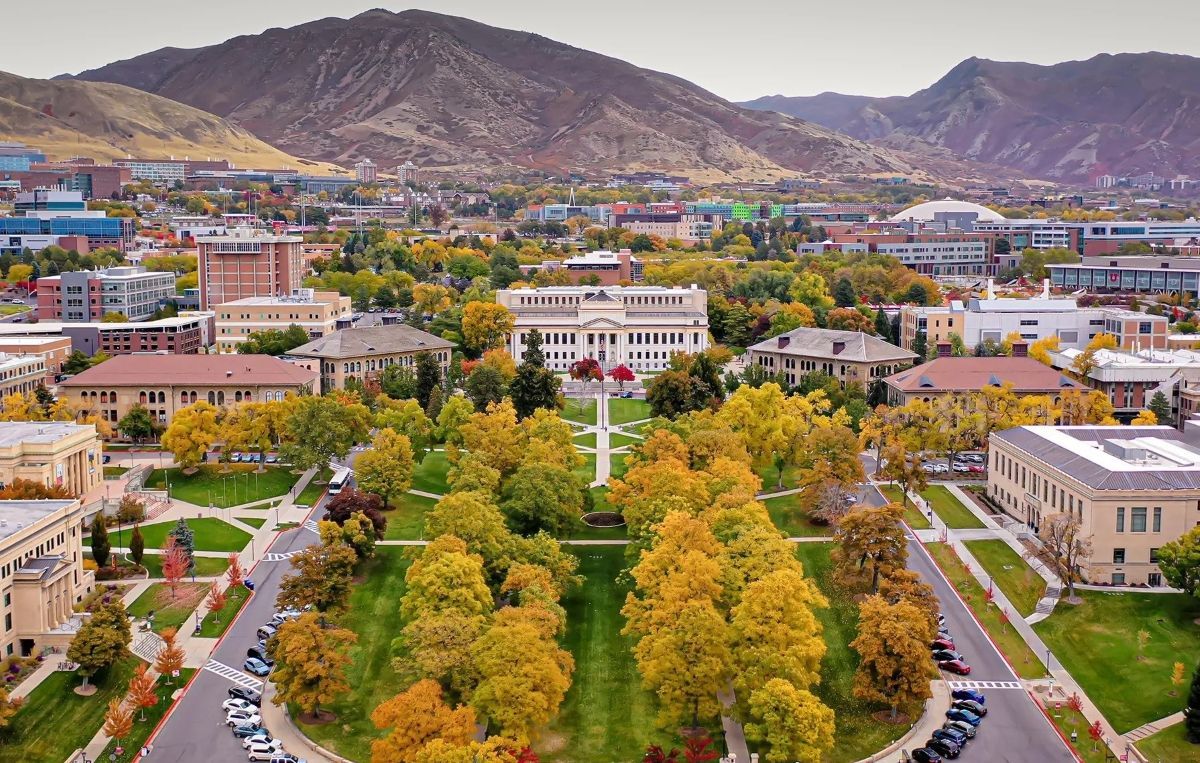 Vibrant autumn colors blanket the University of Utah campus, with academic buildings set against a rugged mountain backdrop under overcast skies.
