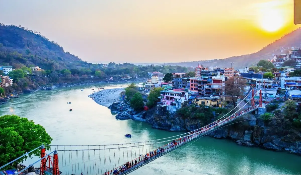 Sunset view of Rishikesh in Uttarakhand, India, featuring the Lakshman Jhula bridge over the Ganges River and colorful buildings along the hillside.