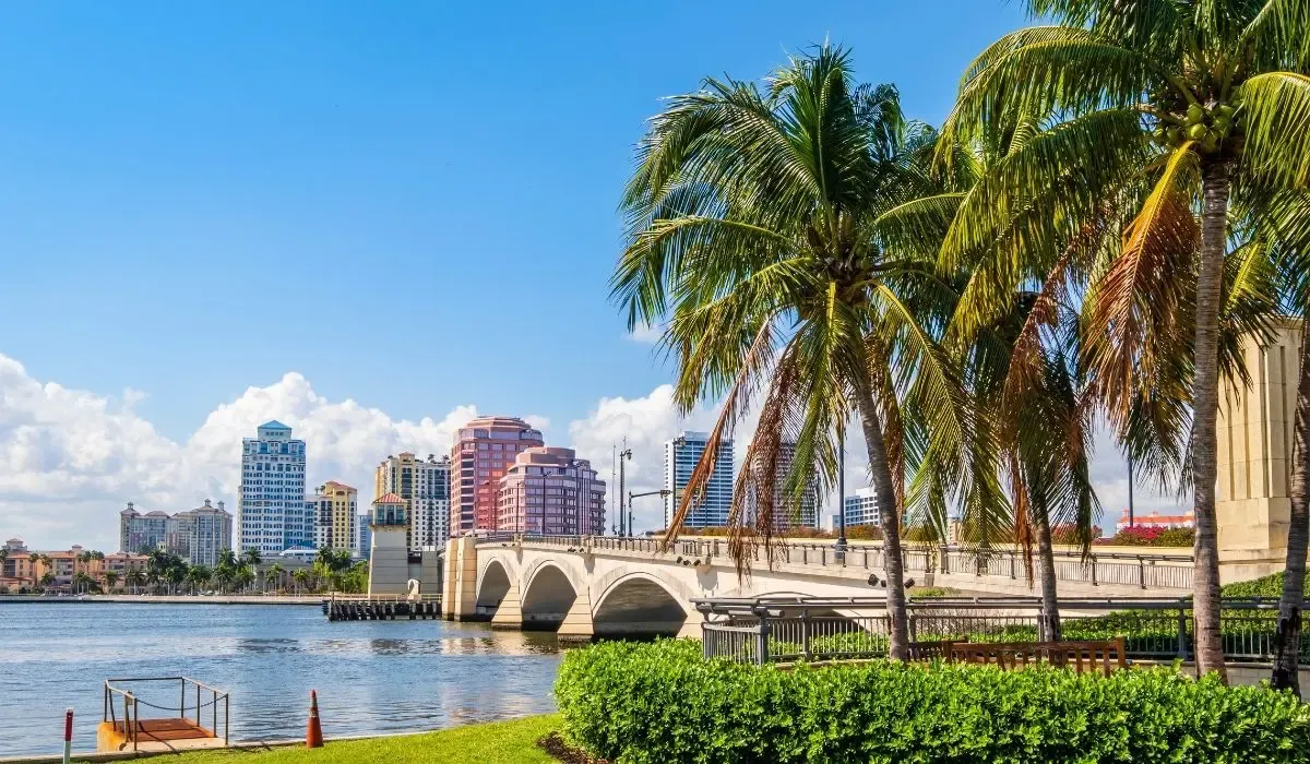 A bustling scene of West Palm Beach's downtown area, highlighting modern high-rise buildings, waterfront parks, and yachts docked along the Intracoastal Waterway.