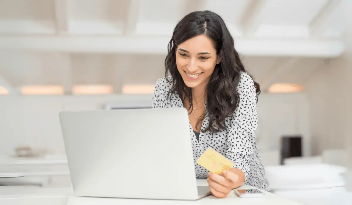 A smiling woman holding a credit card while using a laptop, sitting in a bright, modern office setting.