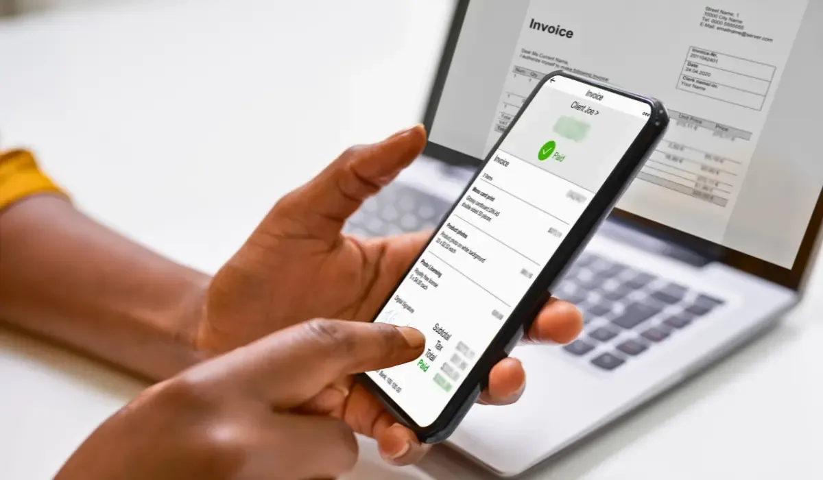A person using a smartphone to check an invoice payment status while sitting at a laptop, with a digital invoice marked as "Paid" on the phone screen.