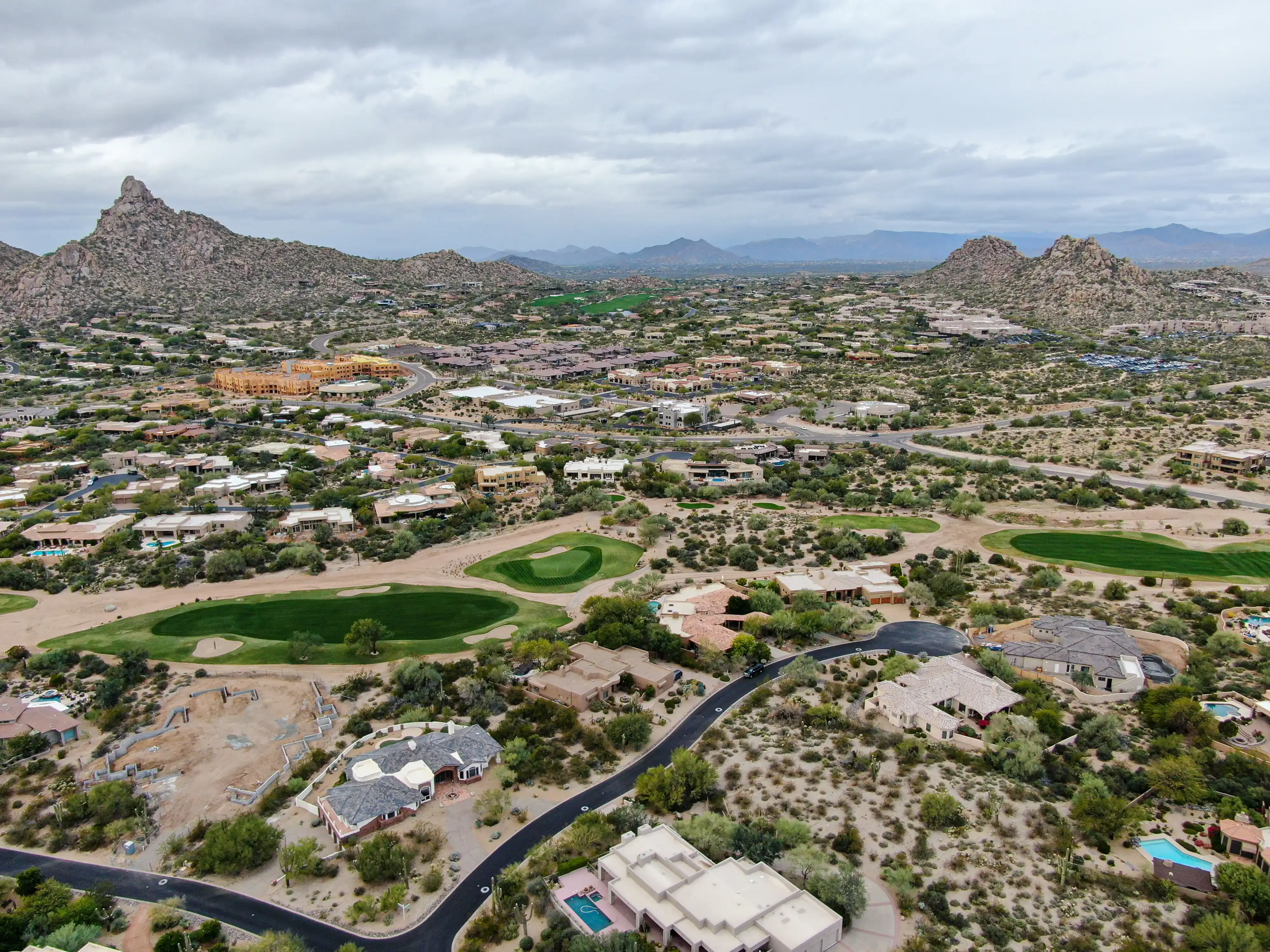 Aerial view of a desert residential community with golf course greens, luxury homes, rocky mountain peaks, and expansive arid landscape under cloudy skies.