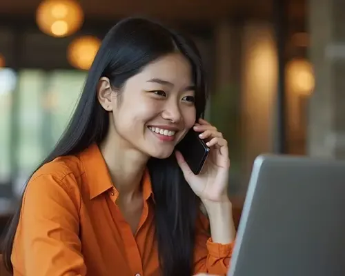 Smiling woman in an orange shirt speaking on the phone and using laptop to send money to Japan.