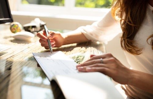 A woman writing a paper cheque at a sunlit desk, illustrating traditional payment methods in contrast to modern digital FX transactions.